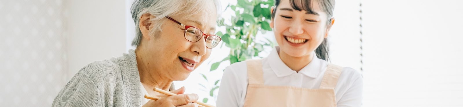 箸を手に、笑顔で食事をする高齢の女性と、隣で笑う職員