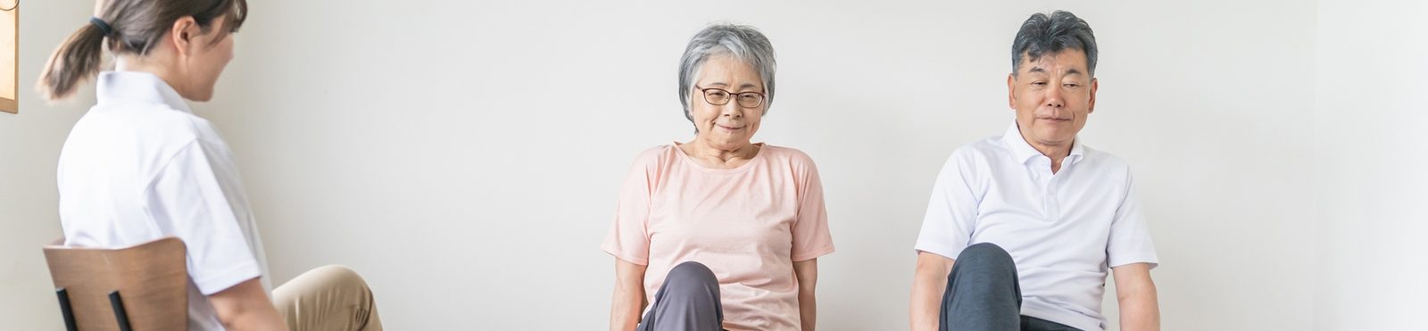 椅子に座って片足を上げる体操をする高齢の男女と、前で見本を見せる指導員