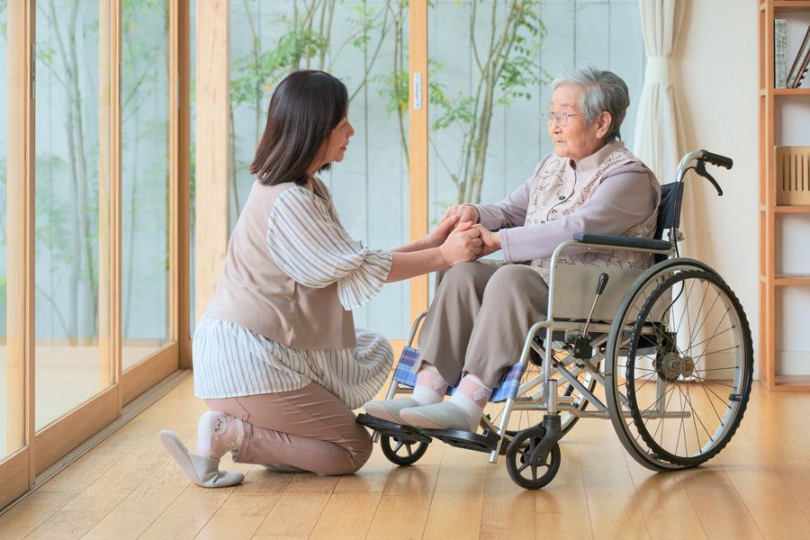 車いすに座る高齢の女性の手を、そばにしゃがんで握る女性