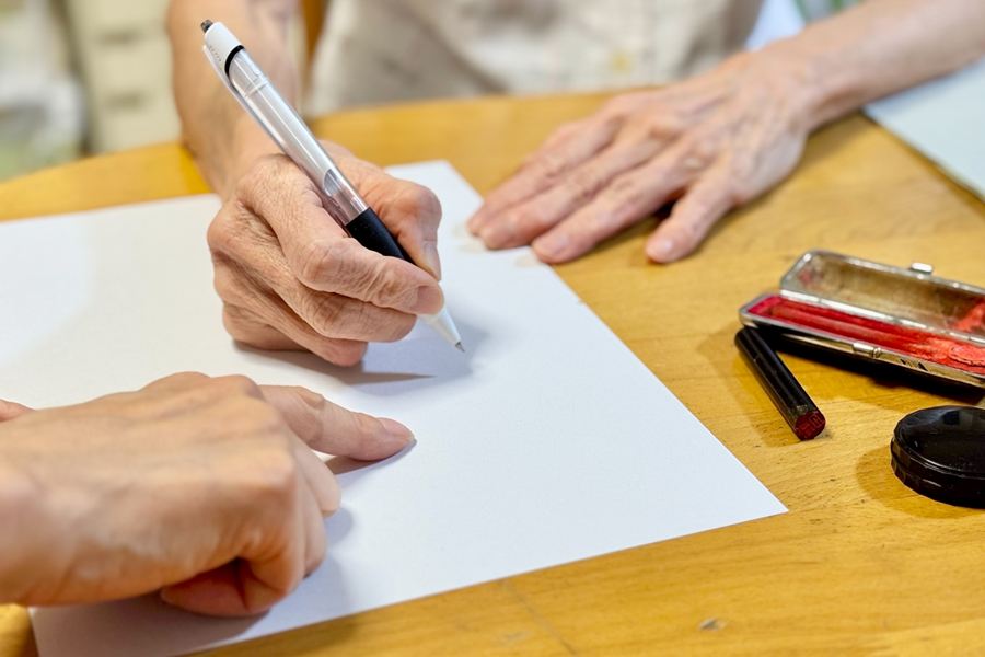 申込書に記入する高齢の女性と、記入する箇所を示すスタッフの手元
