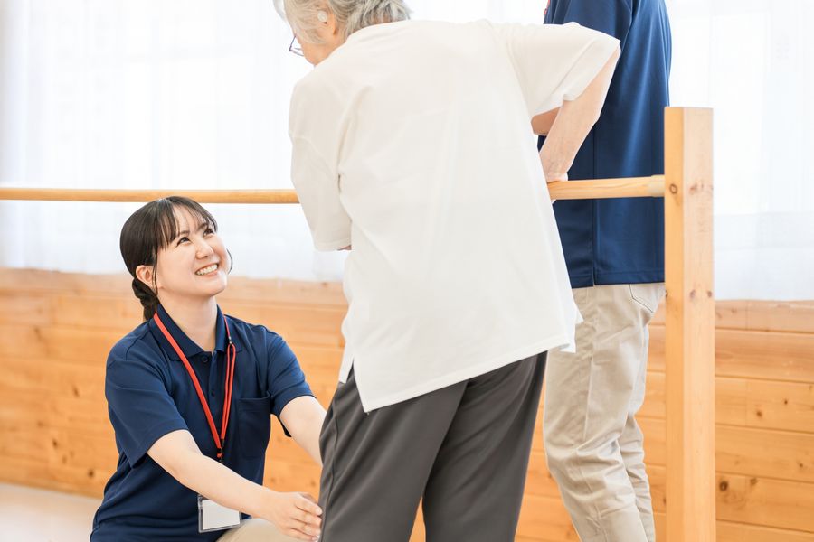 平行棒で歩く練習をする高齢の女性と、しゃがんで笑顔で声をかけるスタッフ