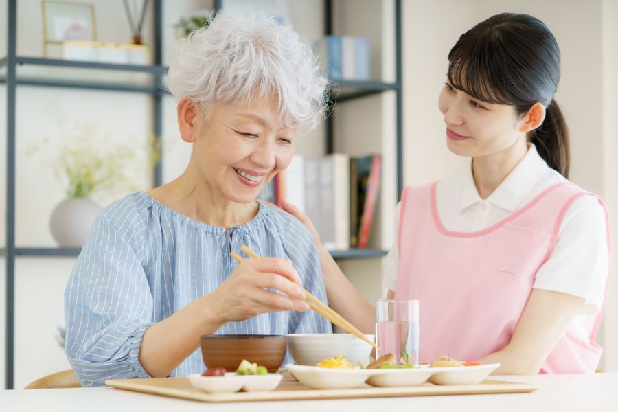 食卓で、箸を手に食事をする高齢の女性と、そばで話しかける職員