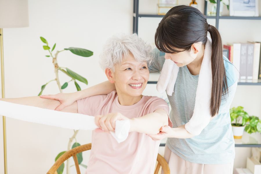 椅子に座ってタオルを両手で持ち、腕を伸ばす高齢の女性と、うしろで支える職員