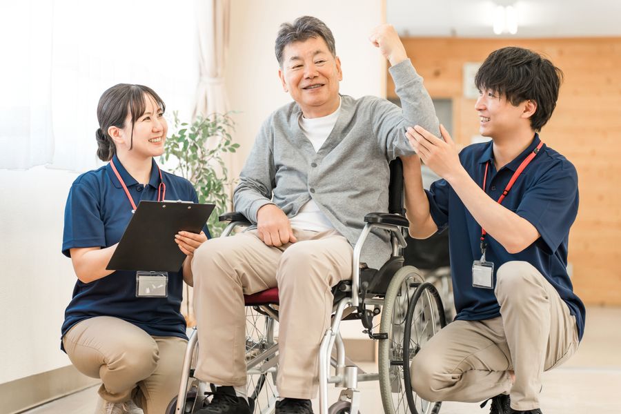 椅子に座って腕を上げる高齢の男性と、そばで声をかける2人の職員