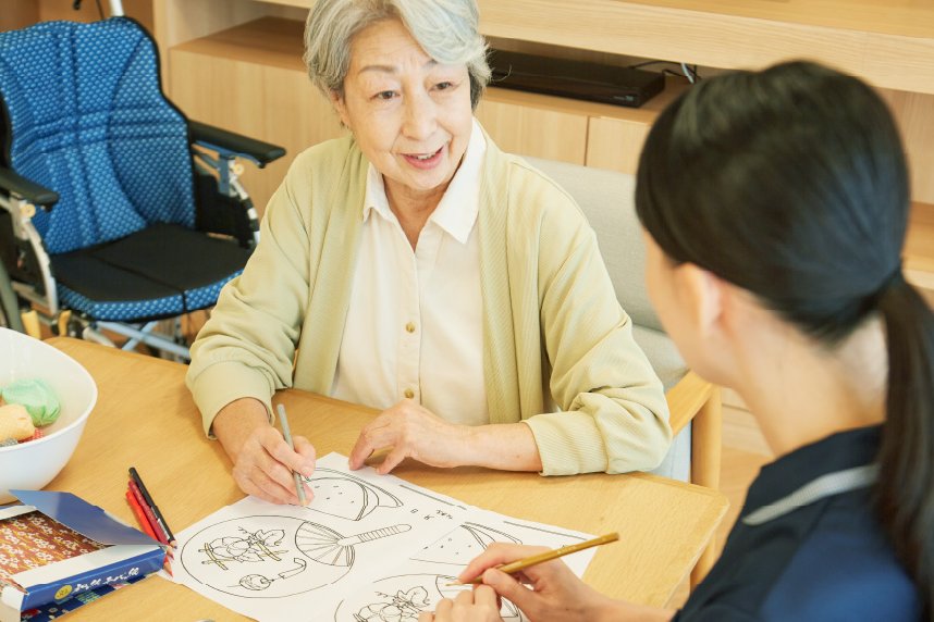 色鉛筆で塗り絵をする高齢の女性と、向かいに座るスタッフ